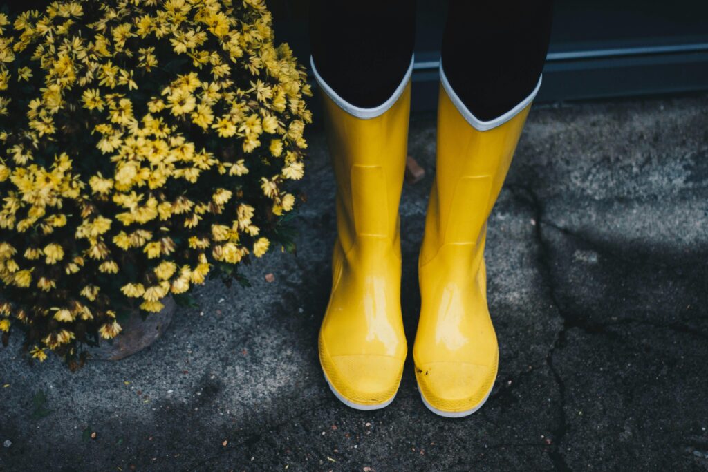 Vibrant yellow rain boots next to blooming yellow flowers on a concrete surface.